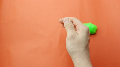 Soft and sticky green slime sits in right hand during ASMR play, its malleable form glowing under light with smooth glossy texture, orange backdrop, static camera, close-up framing