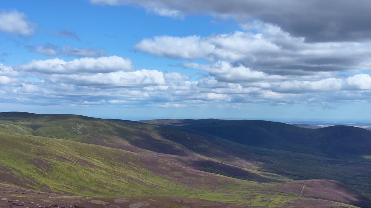Drone pans across sunlit Scottish Highlands, revealing rolling heather hills and dramatic cloudscape
