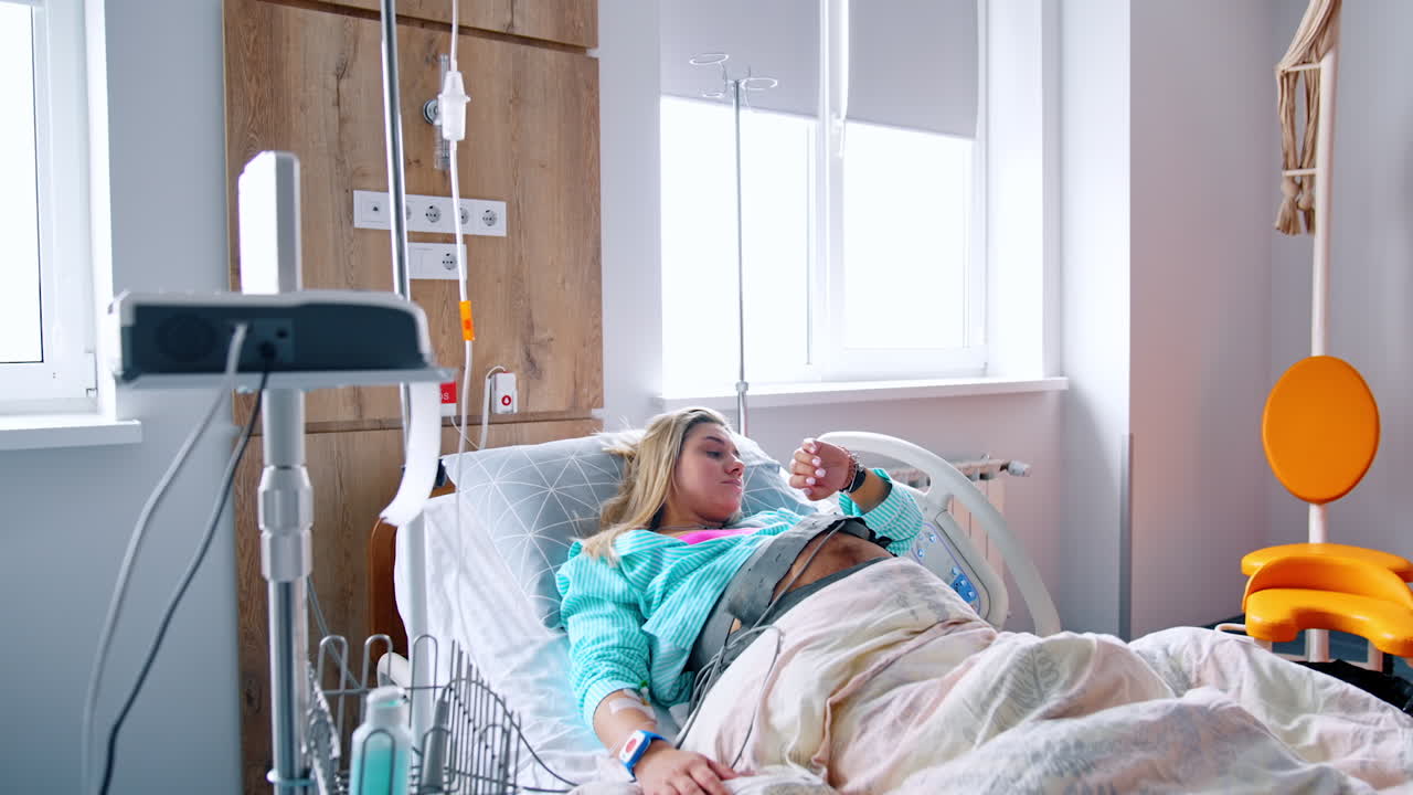 Pregnant blonde Caucasian woman lies in the hospital bed connected to equipment. Cardiotocography belt is on the belly of patient.