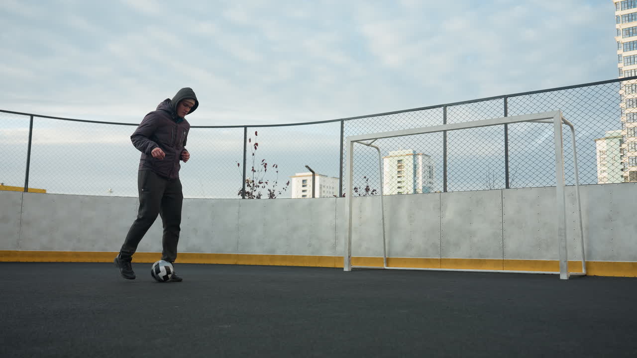 Man practicing soccer drills on sport arena, skillfully controlling ball while focusing on footwork, background includes urban residential buildings, mesh fencing, and goalpost under soft daylight