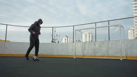 man practicing soccer drills on sport arena, skillfully controlling ball while focusing on footwork, background includes urban residential buildings, mesh fencing, and goalpost under soft daylight