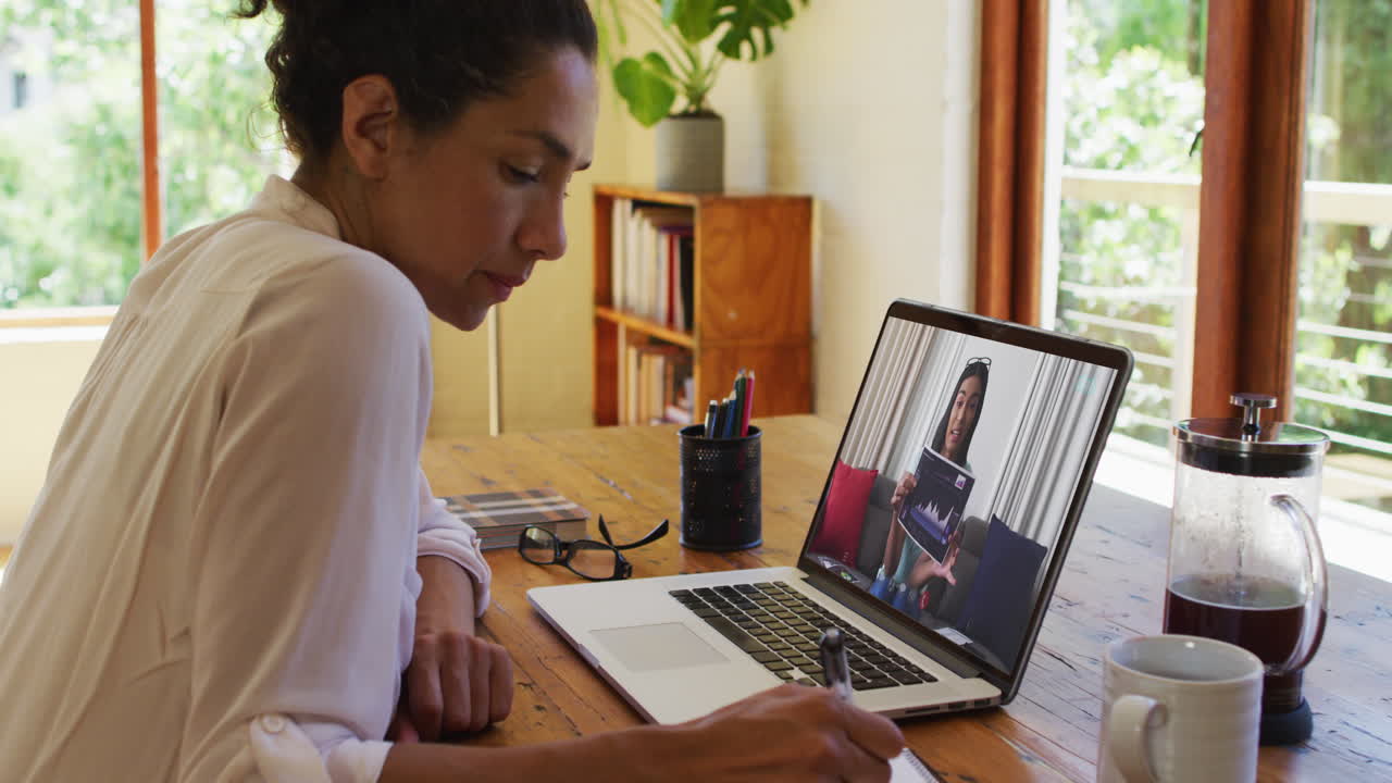 African american woman taking notes while having video call with female colleague on laptop at home