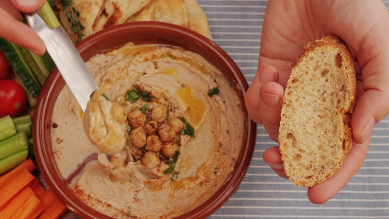 Spreading hummus on bread top view. Hummus garnished with chickpeas, olive oil, spices and greens among fresh vegetables. Humus or fava, a hearty snack of legumes. Kosher healthy vegan food. Lean, vegetarian dish