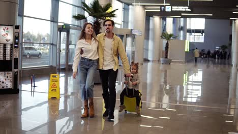 Portrait-of-smilling,-modern-caucasian-family---mother,-father,-daughter---walking-the-airport-hall-and-riding-their-daughter-on-suitcase-before-the-departure-to-the-vacation.-Slow-motion