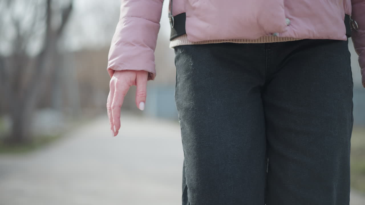 Close-up view of woman walking outdoors in winter, showing hand and torso in pink jacket and black pants, with soft sunlight and blurred background of park trees and paved path during cold season