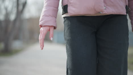 close-up view of woman walking outdoors in winter, showing hand and torso in pink jacket and black pants, with soft sunlight and blurred background of park trees and paved path during cold season