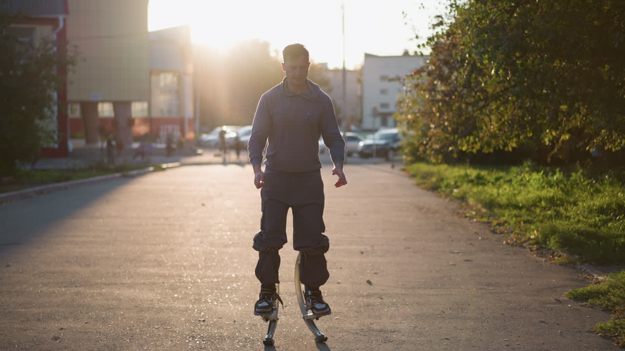Front shot of man jumping on stilts wearing gray hoodie and dark gray pants during sunset in residential area surrounded by houses and people, showcasing athletic energy, balance, motion