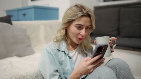 smiling girl is messaging online on the phone, holds mug with hot drink, checks emails