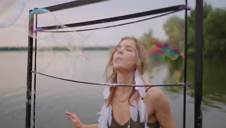 a young female artist blows a lot of soap bubbles shows a theatrical show using a frame in slow motion at sunset on a lake