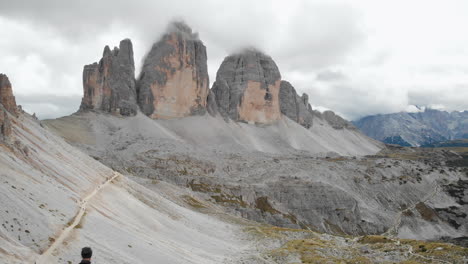 Aerial-shot-from-a-drone-passing-by-a-male-model-with-backpack-looking-at-Tre-Cime-di-Lavaredo-in-the-Dolomites,-Italy