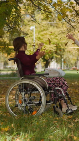 mother with legs injury and daughter play in park. girl throws up yellow leaves handful and couple waves hands looking upward joyfully. loving family on vacation