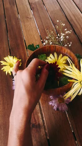 a hand holding a flower over a bowl of flowers on a wooden table