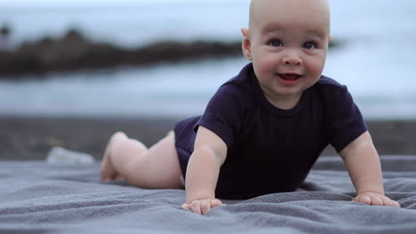 on the black sand by the ocean, the baby lies on his stomach, laughing as he gazes at the camera