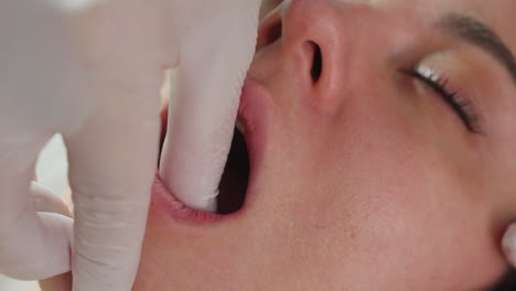 Close up of young woman wrapped in clinical blanket closing eyes and opening mouth relaxed manner as skin care expert wearing gloves dips finger tip into mouth rubbing jaw line gently
