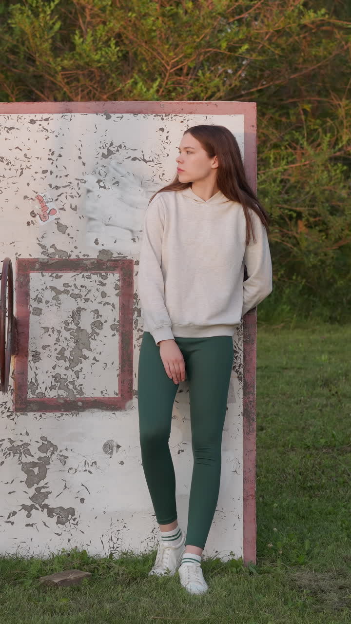 Woman leans on old basketball backboard. Lonely woman in sportswear stands among green grass on abandoned basketball court. Encapsulating moment of solitude