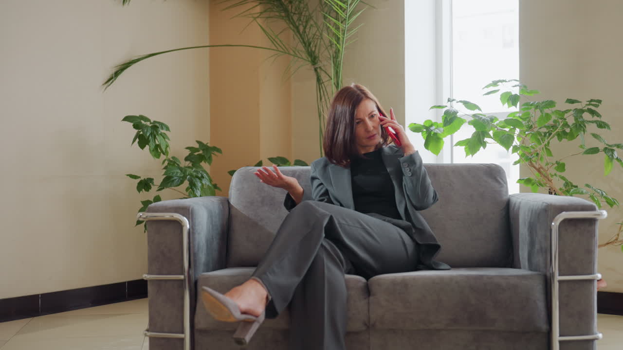 Confident businesswoman talking smiling on red phone while sitting cross-legged on grey sofa in bright modern office with indoor plants and sunlight streaming through large window