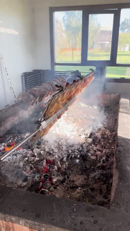 vertical handheld video of a rotating grill with numerous chicken legs roasting over charcoal. glowing red coals and white smoke fill the rustic indoor setting, with a green outdoor view..