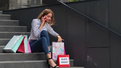 girl sitting on stairs with bags talking on smartphone about sale in shopping mall in black friday