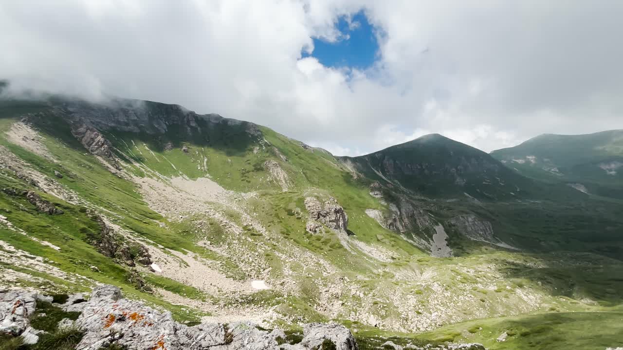 A mountain landscape featuring rocky slopes, grassy areas under a cloudy sky. The mountain peaks are partly obscured by clouds, with sunlight breaking through in spots.