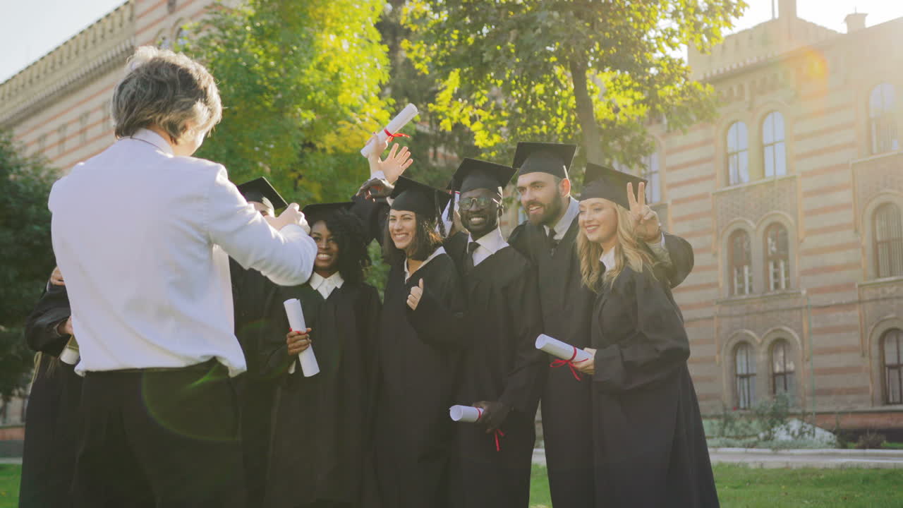 Back view on the gray-haired man taking a photo portrait with the smartphone of the multi ethnical graduates in traditional black clothes and caps with diplomas in hands