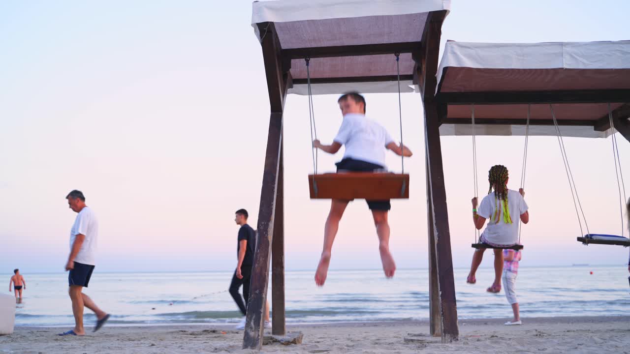 Children on a swing against the sea. Back view of a boy and girl are swinging on a playground on beach in the evening in summer.