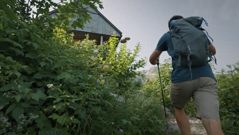 camera tracking a young hiker walking up a path towards the wooden cottage surrounden with green plants