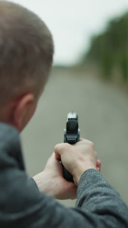 una vista de cerca desde atrás de un hombre en una chaqueta gris apuntando una pistola por una vía ferroviaria desierta, con un fondo de bosque borroso