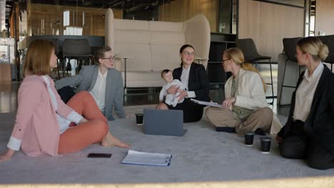 a group of businesswoman in business clothes sit in a circle on a gray carpet and communicate while a brunette girl in a business suit holds her little baby daughter in her arms in a modern office