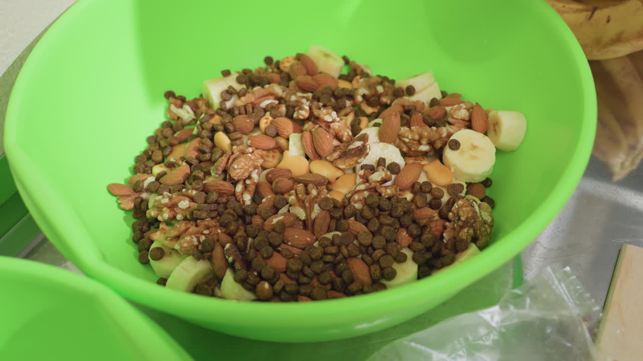 green bowl filled with kibble and toppings closeup of crunchy brown kibble mixed with sliced banana, almonds, oats and seeds in bright green plastic bowl on kitchen countertop, natural light