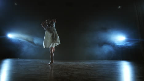 a young girl in a white dress dances a modern ballet makes rotations and jumps in slow motion on the stage with smoke against the spotlights in full shot.