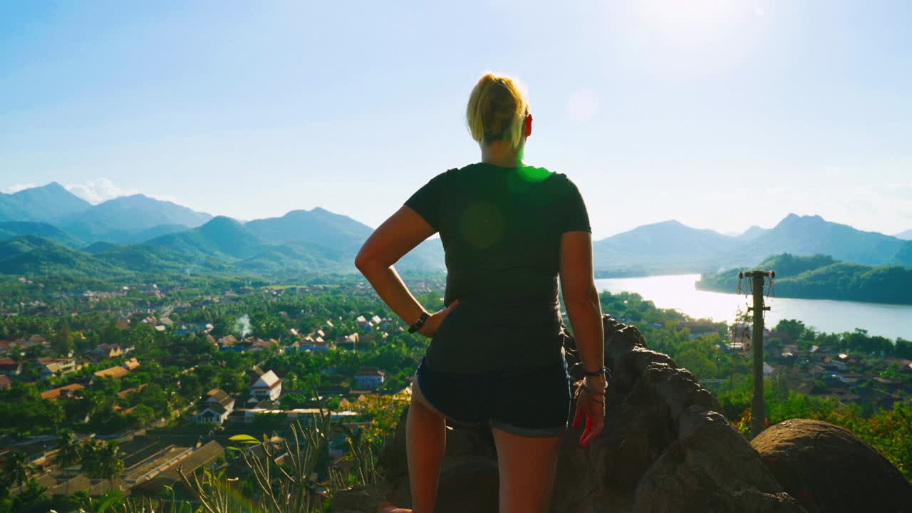 joven caucásica de pie en la cima de un acantilado disfrutando de la impresionante vista de luang prabang y el vasto paisaje verde del sudeste asiático