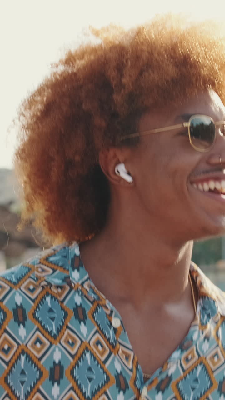 Giovane uomo felice con capelli ricci e occhiali da sole che sorride all'aperto
