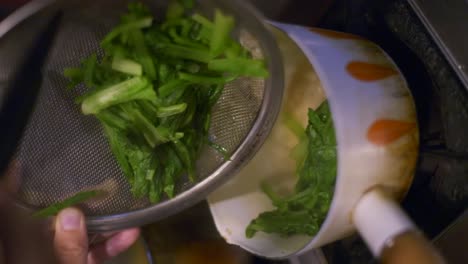 freshly cooked green vegetables moved from metal strainer into small white cooking pot, filmed as vertical slow motion closeup shot in handheld style