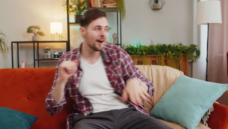 Man-singing-and-dancing-celebrate-victory-win-enjoying-music-sitting-on-couch-in-living-home-room