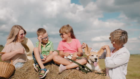 la familia sonriente disfruta del campo mientras la abuela juega con orejas de perro, los niños se relajan en el pajar, la mujer con la canasta se ríe, el campo iluminado por el sol con balas de heno y el cielo azul