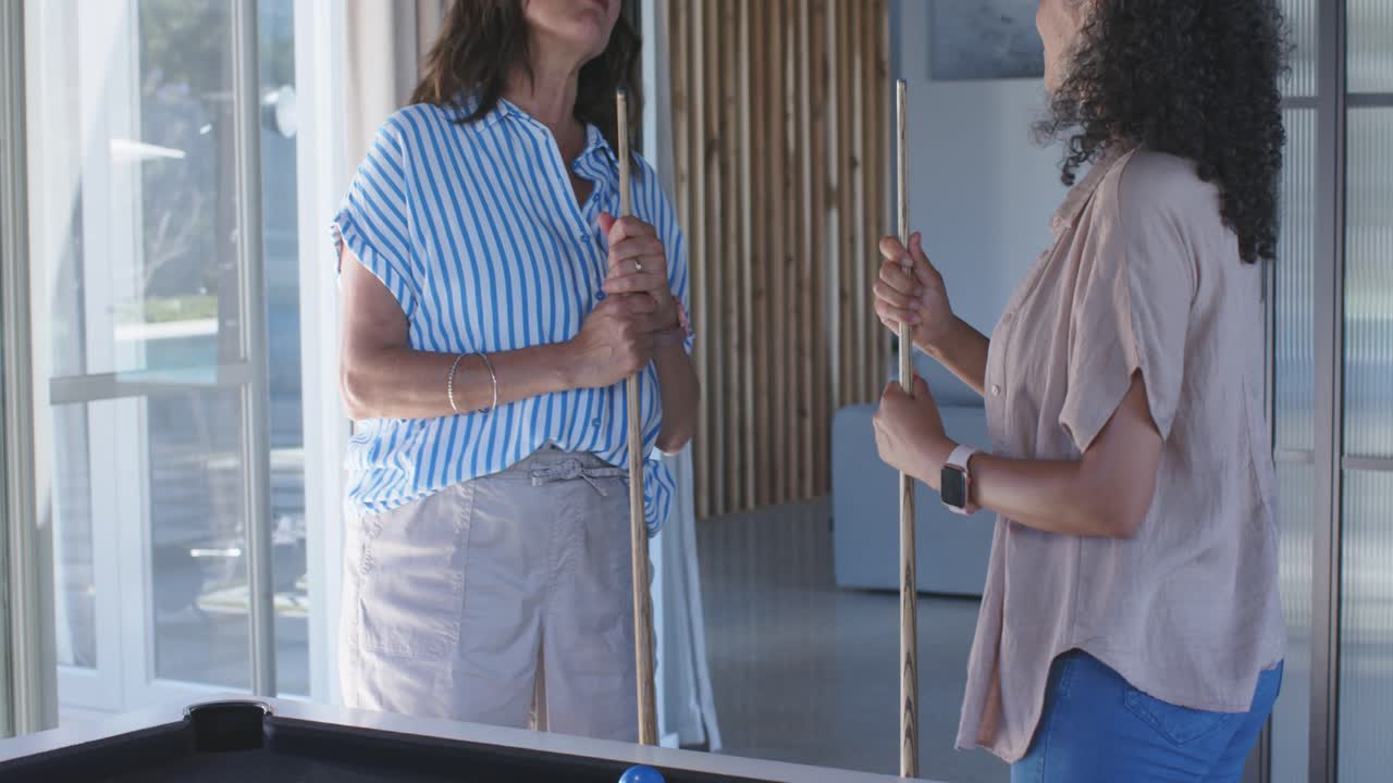 Talking women in striped blouse and beige top holding pool cues at home near pool table