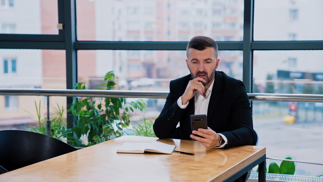 Serious man wearing suit sits at desk talking and holding phone. Boss searching for information in internet on the smartphone.