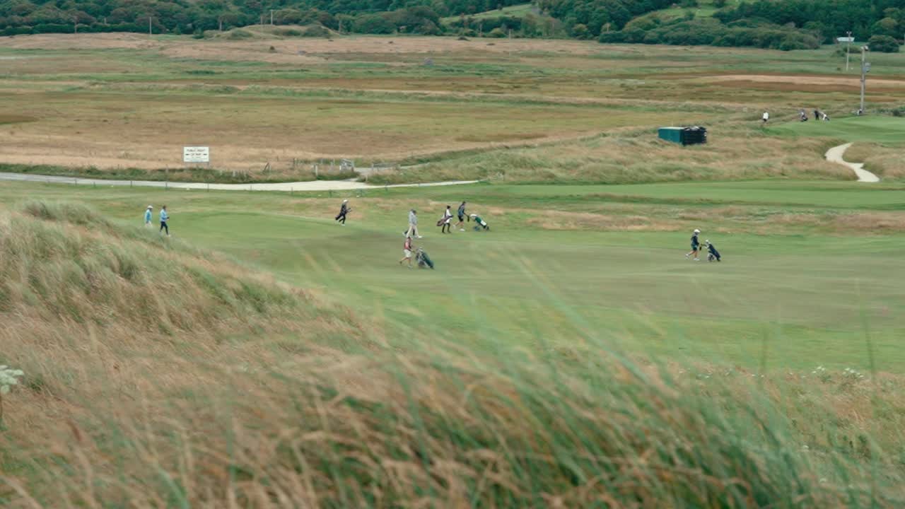 골퍼들과 그들의 캐디들이 푸시 카트를 사용하여 페어웨이를 가로질러 고 있으며, 높은 잔디에서 느린 움직임으로 바람에 부히는 모습입니다.