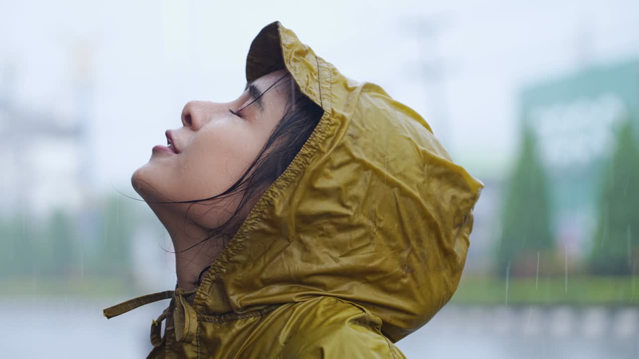노란색 레인코트를 입은 어린 아시안 소녀가 도로 에 서서 하늘을 바라보고 얼굴에 비가 내리는 것을 즐기고 미소 짓고, 비가 오는 계절, 날씨, 기후, 비가 내리고, 신선함을 느다.