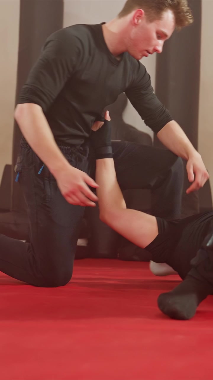 Two young men practice martial arts indoors on red mat. One man in black attire dominates other prone men. Close up shots emphasize arm interactions while wider views show