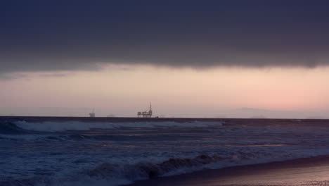 oil rigs in the distance at sun set off the coast as waves crash on the beach