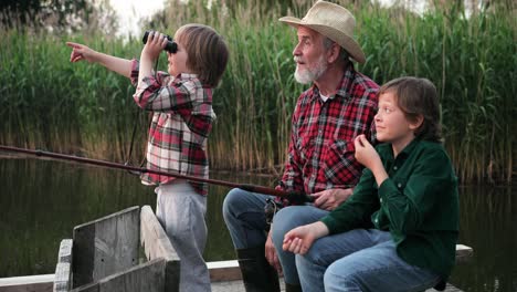 cute little boy using binoculars and showing something to his grandfather and his brother while they are fishing sitting on the lake pier