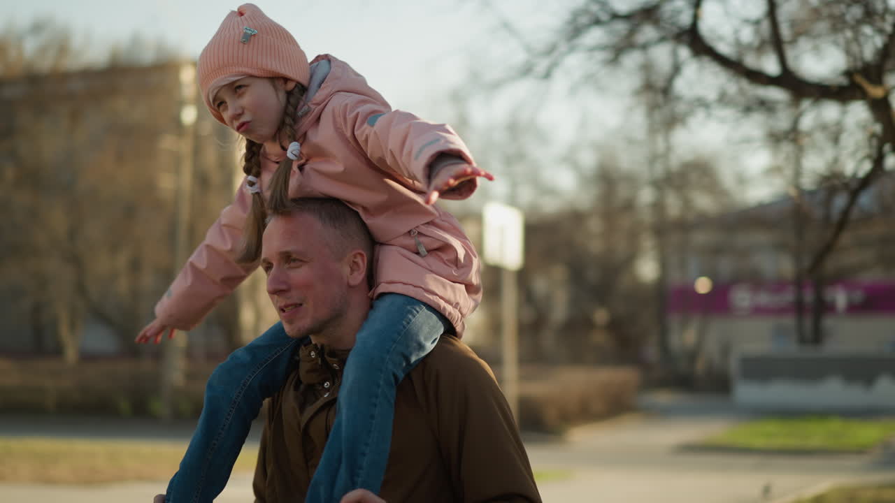Little girl in a pink cap and jacket joyfully spreads her arms while sitting on her father's neck as the camera circles around them
