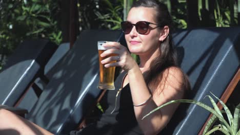 Plano-Lateral-Medio-De-Una-Mujer-Joven-Disfrutando-De-Un-Vaso-De-Cerveza-En-Una-Tumbona-Junto-A-Una-Piscina-Tropical