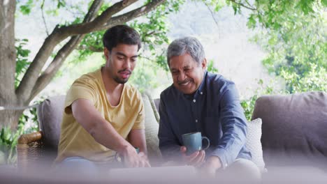 Slow-motion-shot-of-smiling-biracial-father-and-son-holding-cups-while-discussing-over-laptop