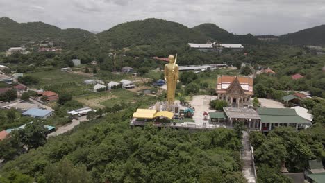 Aerial-view-of-Buddhist-temple-at-lush-Hilltop,-Standing-Golden-Buddha-Statue