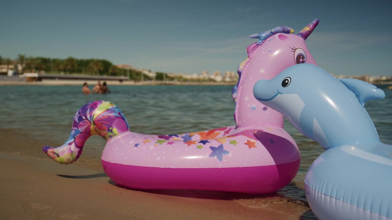 Colorful unicorn and dolphin inflatable floats on the shallow water at Plage de la Gravette with families swimming and the rocky breakwater in the background