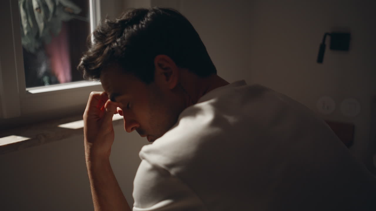 Homem ponderando problemas no quarto perto da janela ao luar. Cara sem sono na cama