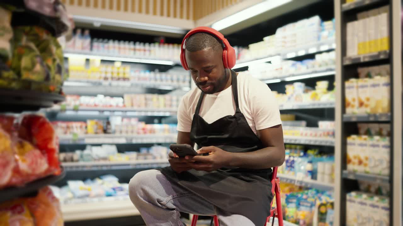 a man with Black skin color in red headphones and a black apron sits on a chair and listens to music in a supermarket. Break at work