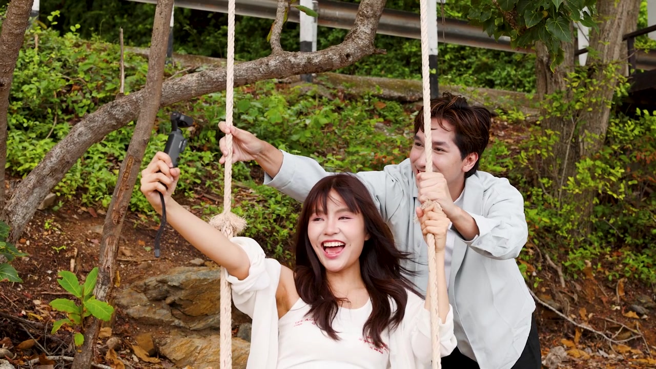 คู่รักชาวเอเชียยิ้มแย้มขณะบันทึกวิดีโอวล็อกบนชิงช้าไม้กลางแจ้ง